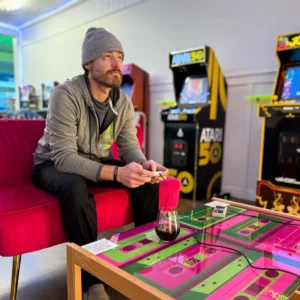 Man playing video games with arcade games in background and glass of wine on the table