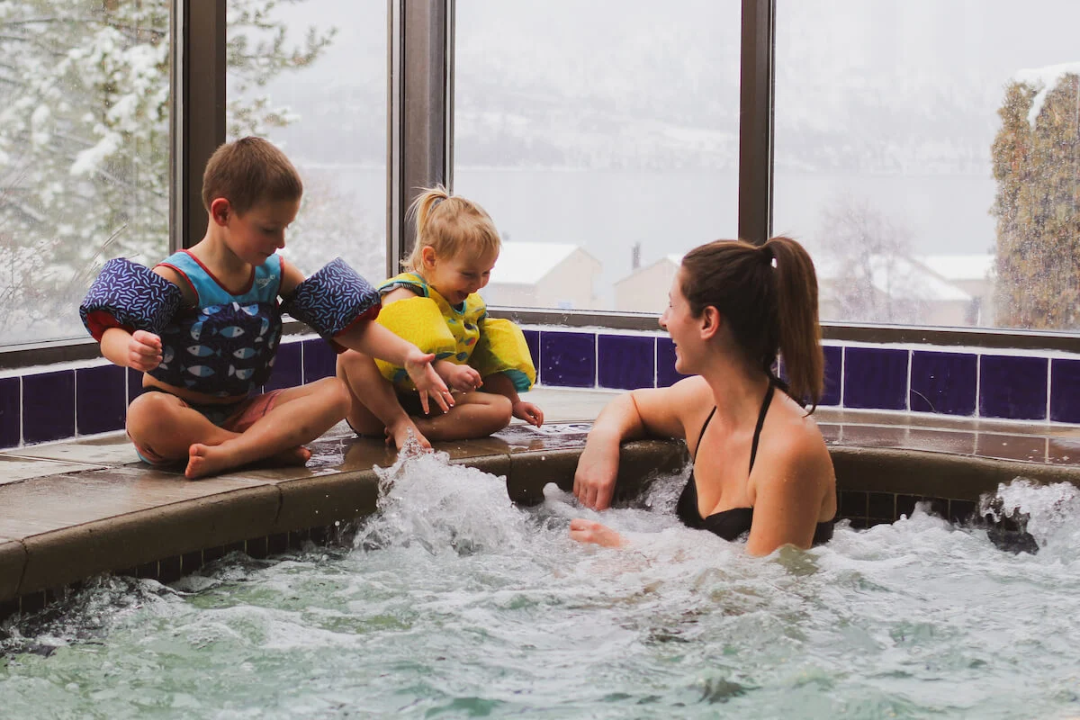 Woman in hot tub with two children playing on the edge with snowy scenery in background